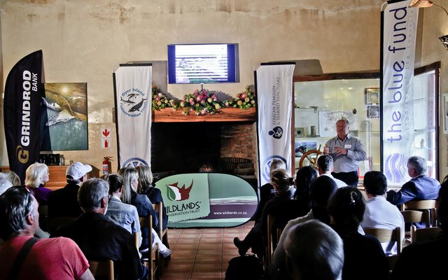 Voor links is Wilfred Chivell, stigter van Dyer Island Conservation Trust, besig om die gaste oor die agtergrond van die beplande African Penguin & Seabird Sanctuary in te lig, kort vóór die sooispit-seremonie